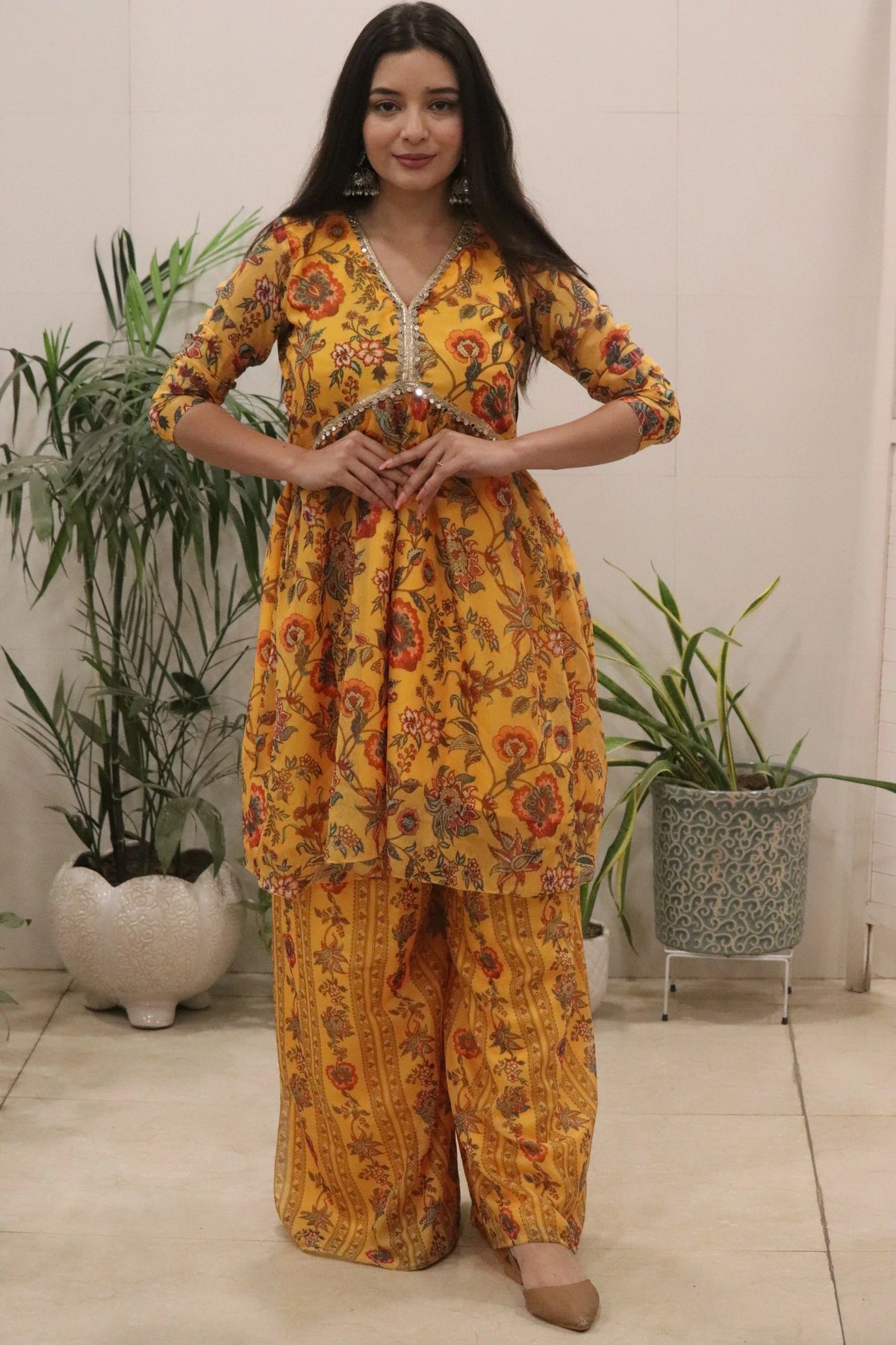 Woman wearing a yellow floral outfit standing indoors with plants in the background
