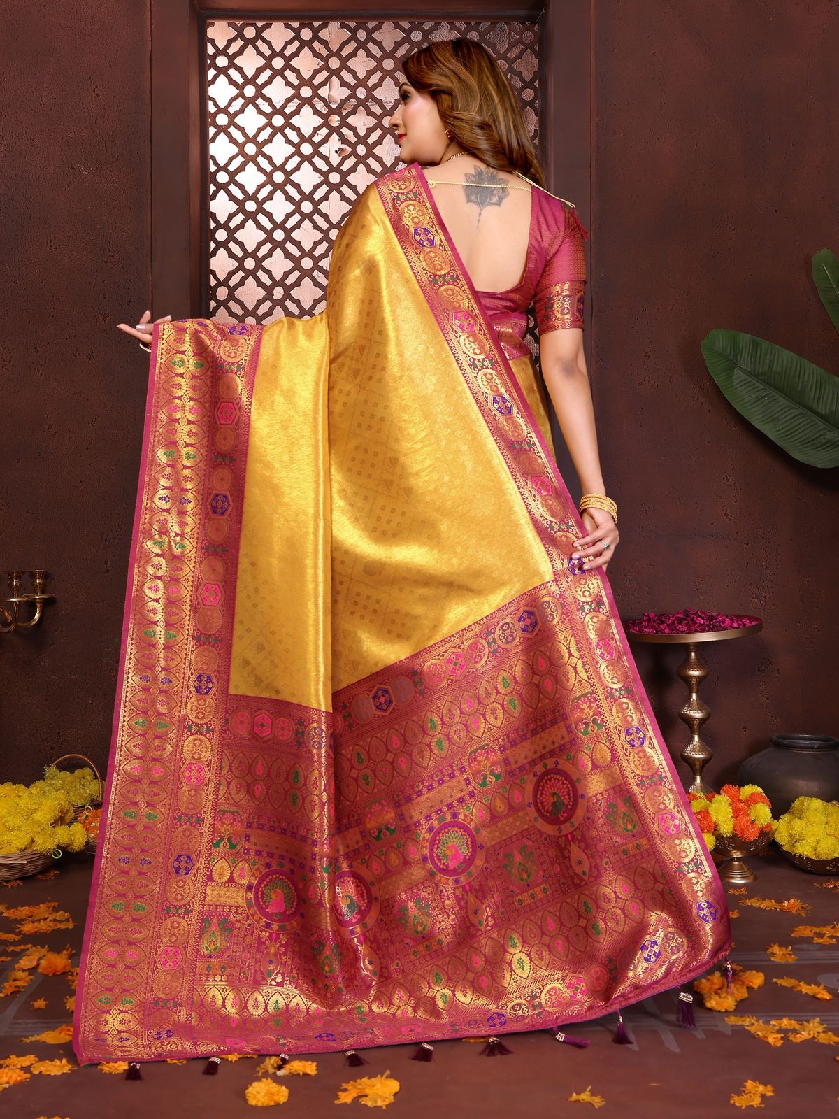 Woman in a traditional pink and yellow saree with intricate patterns, standing against a decorative wall.