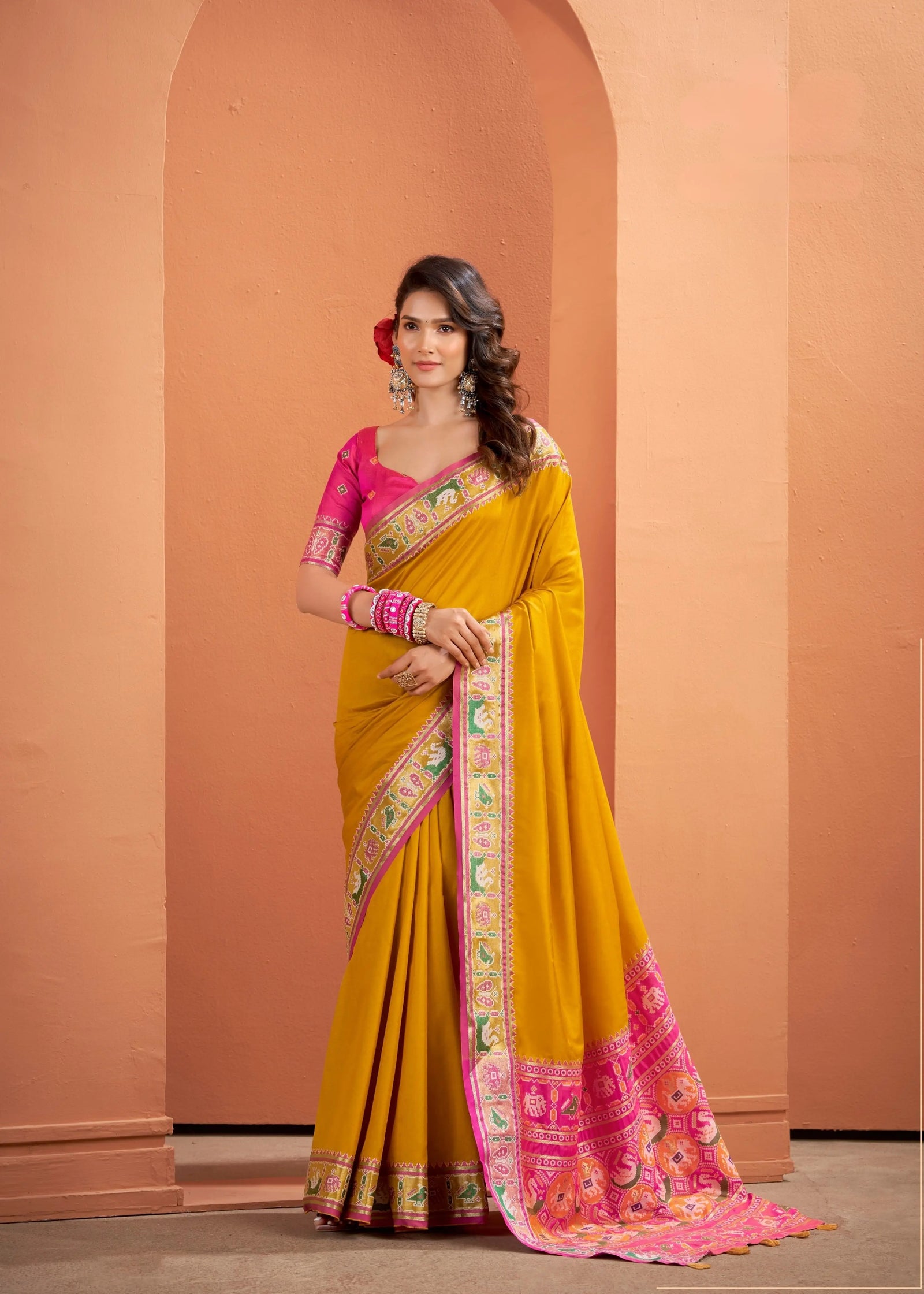 Woman wearing a yellow saree with pink blouse against a peach-colored wall.
