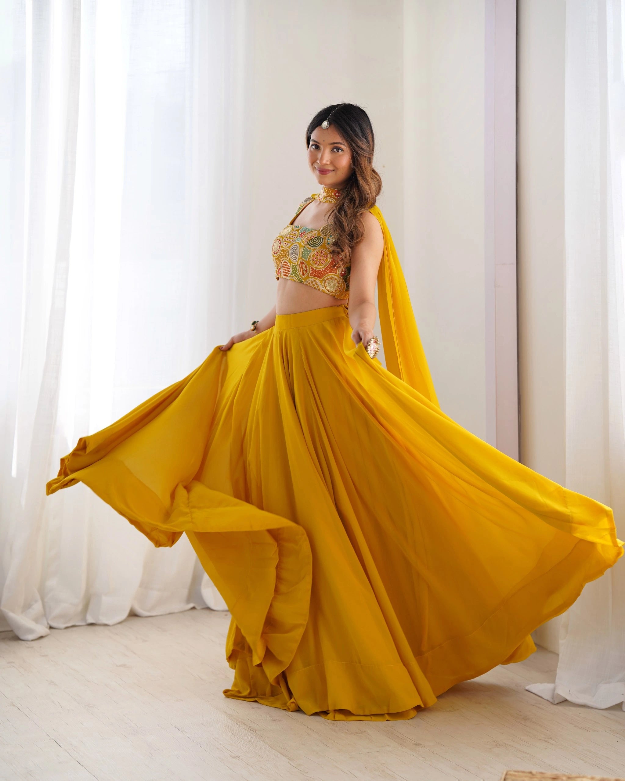 Woman in a yellow traditional outfit with a floral top, standing against white curtains.