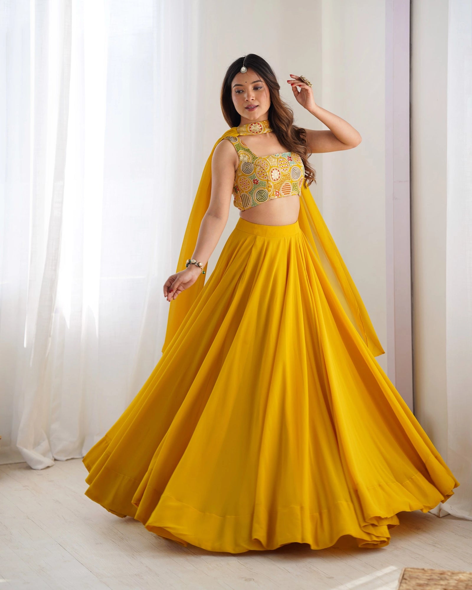 Woman wearing a yellow traditional outfit with a floral top and flowing skirt against a white curtain background.
