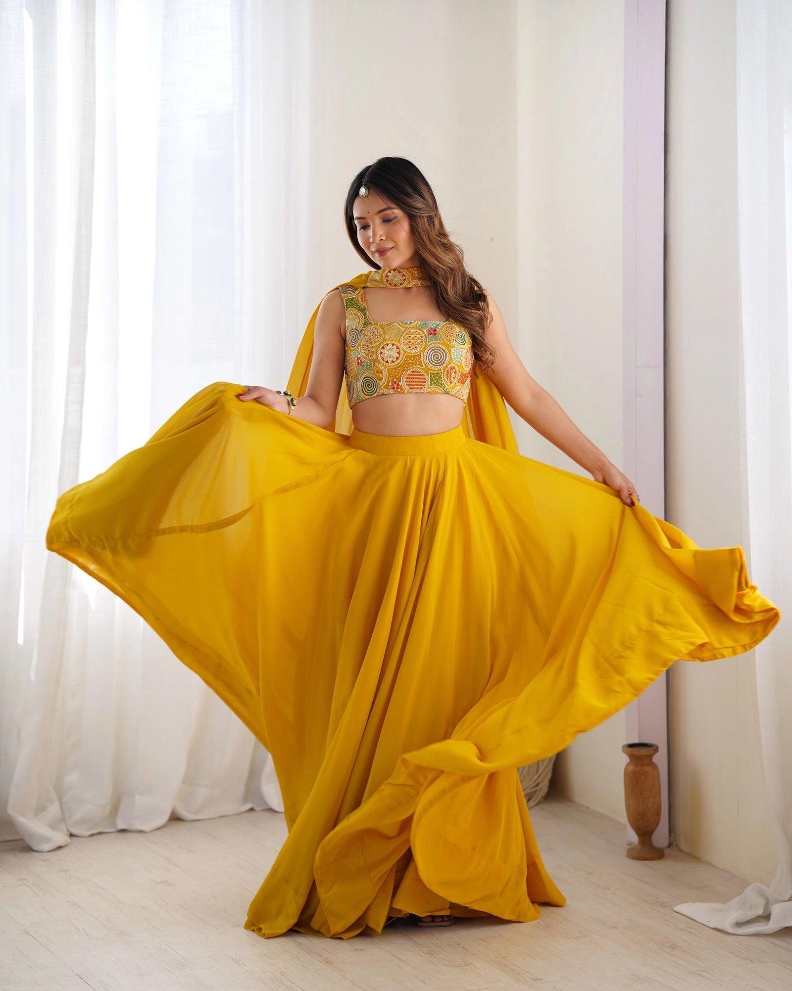 Woman in a yellow traditional outfit with a floral top, standing indoors with white curtains in the background.