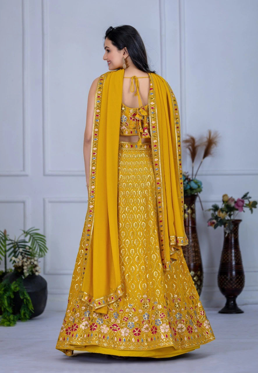 Woman in a yellow embroidered traditional outfit with a matching dupatta, standing against a white paneled wall.
