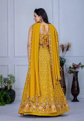 Woman in a yellow embroidered traditional outfit with a matching dupatta, standing against a white paneled wall.