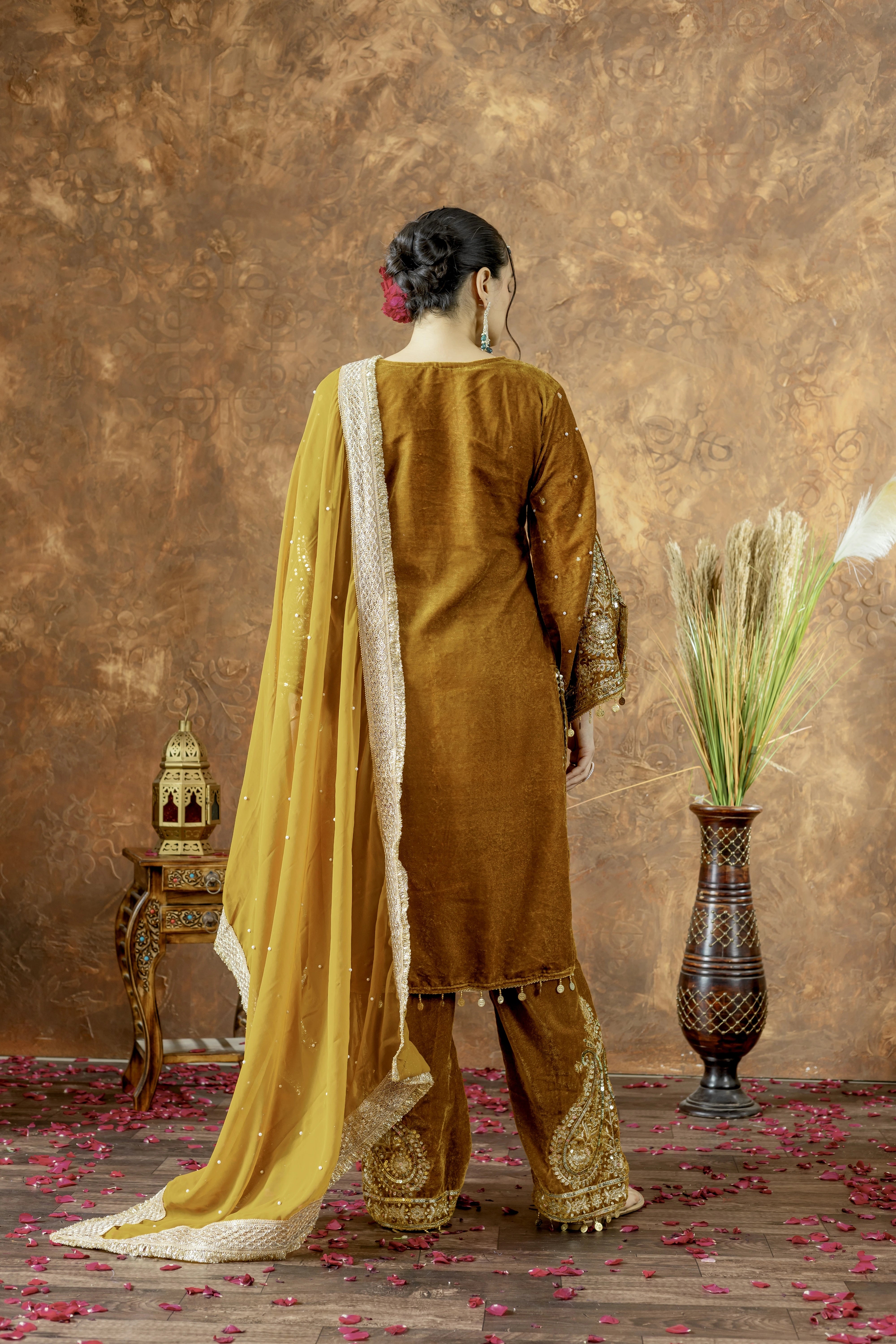 Woman in a mustard yellow traditional outfit with a gold border, standing against a textured brown wall.