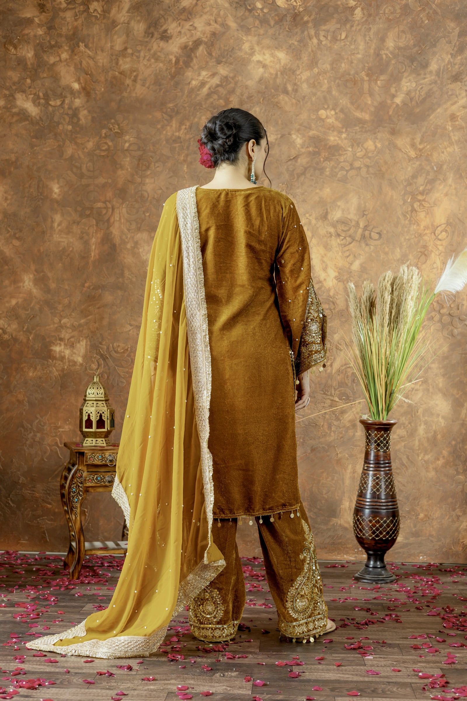 Woman in a mustard yellow traditional outfit with a gold border, standing against a textured brown wall.