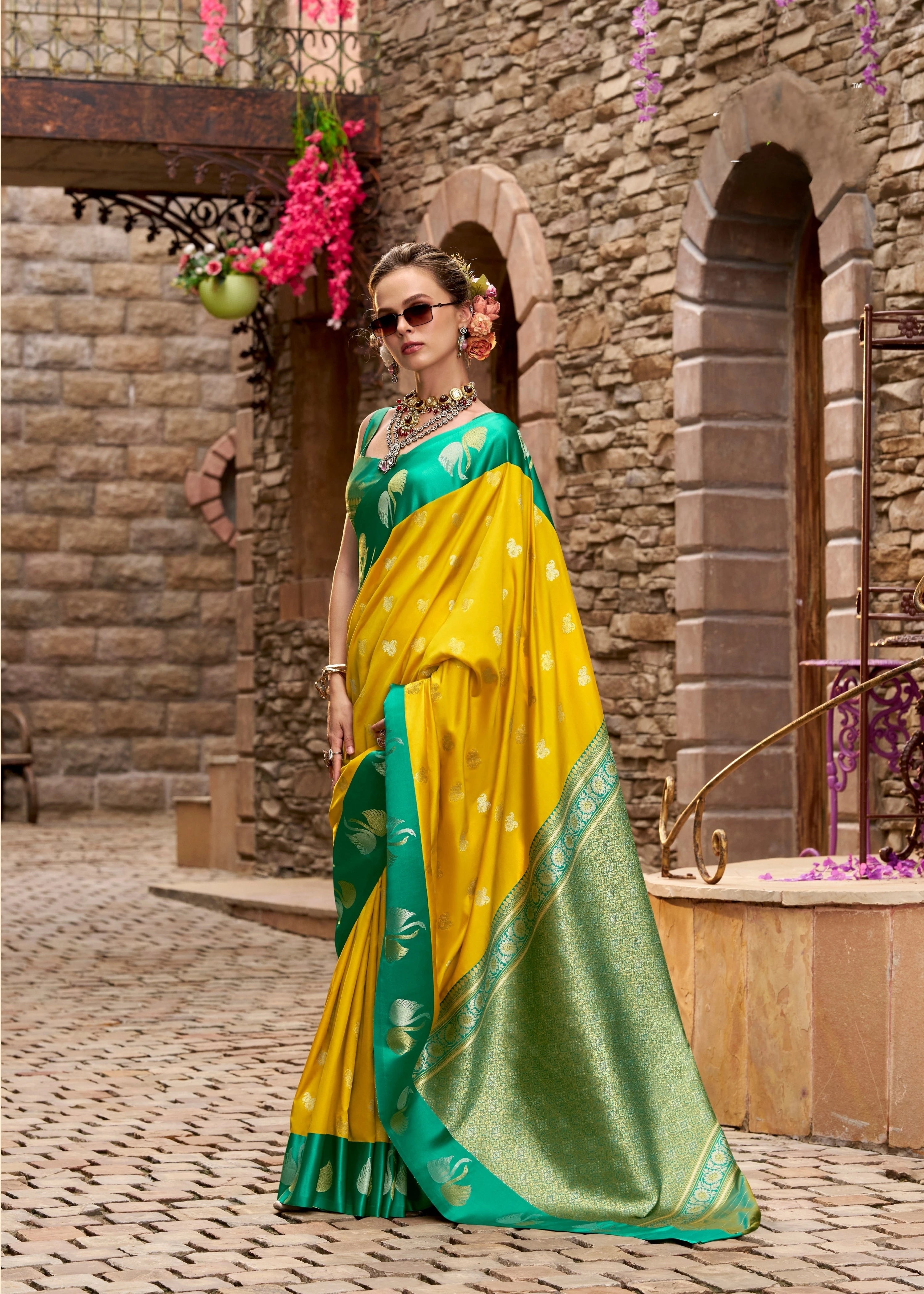 Woman in a yellow and green saree standing in front of a stone wall.