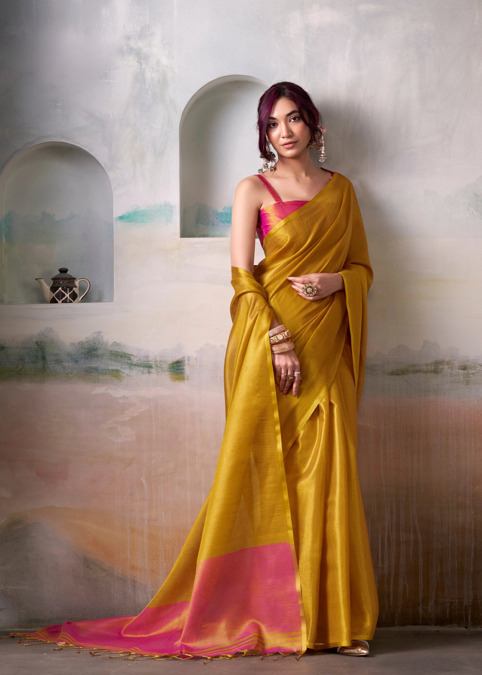 Woman wearing a yellow saree with pink blouse against a textured wall.