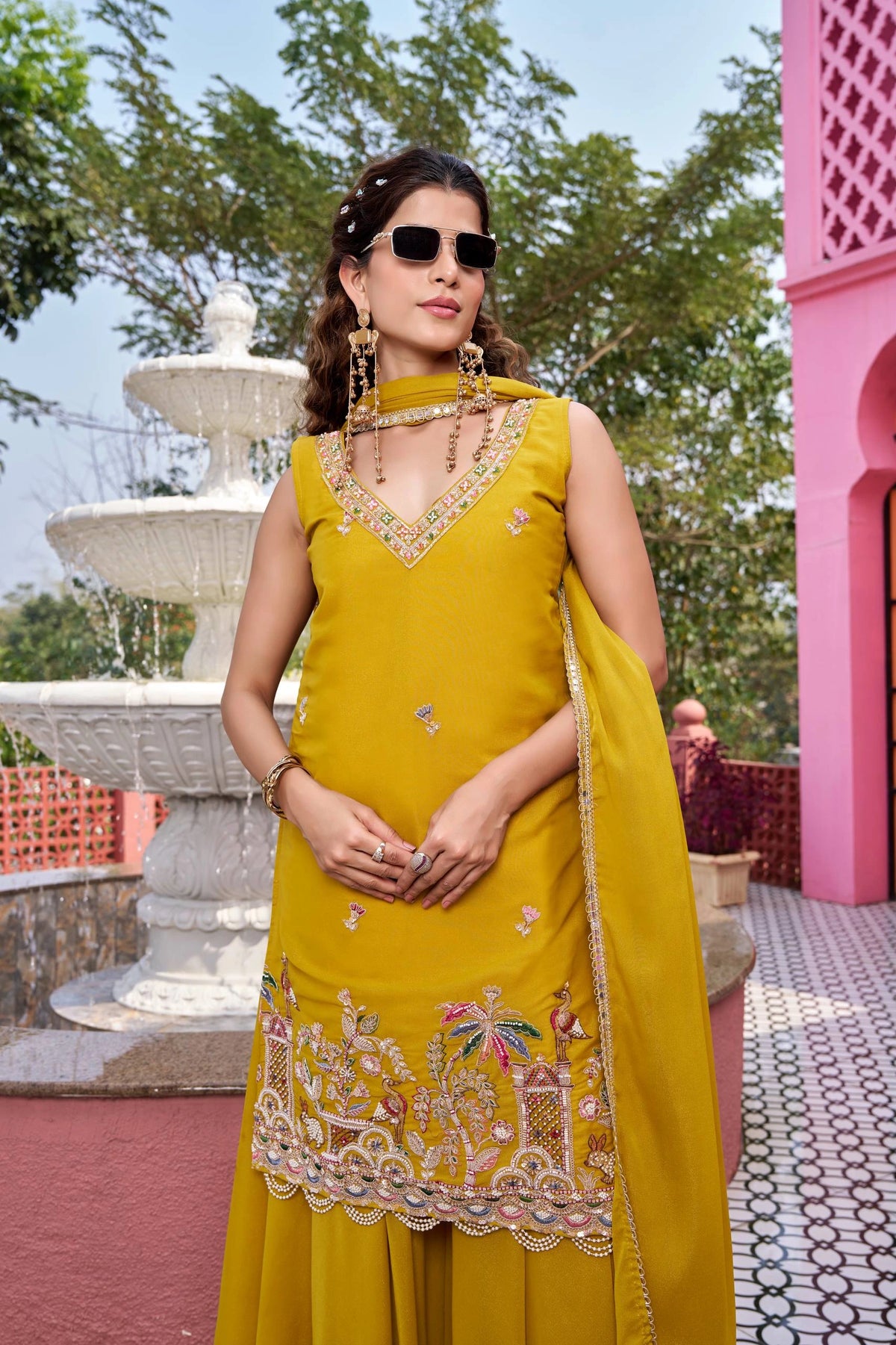 Woman in a yellow traditional outfit with embroidery, standing outdoors near a fountain and pink structure.