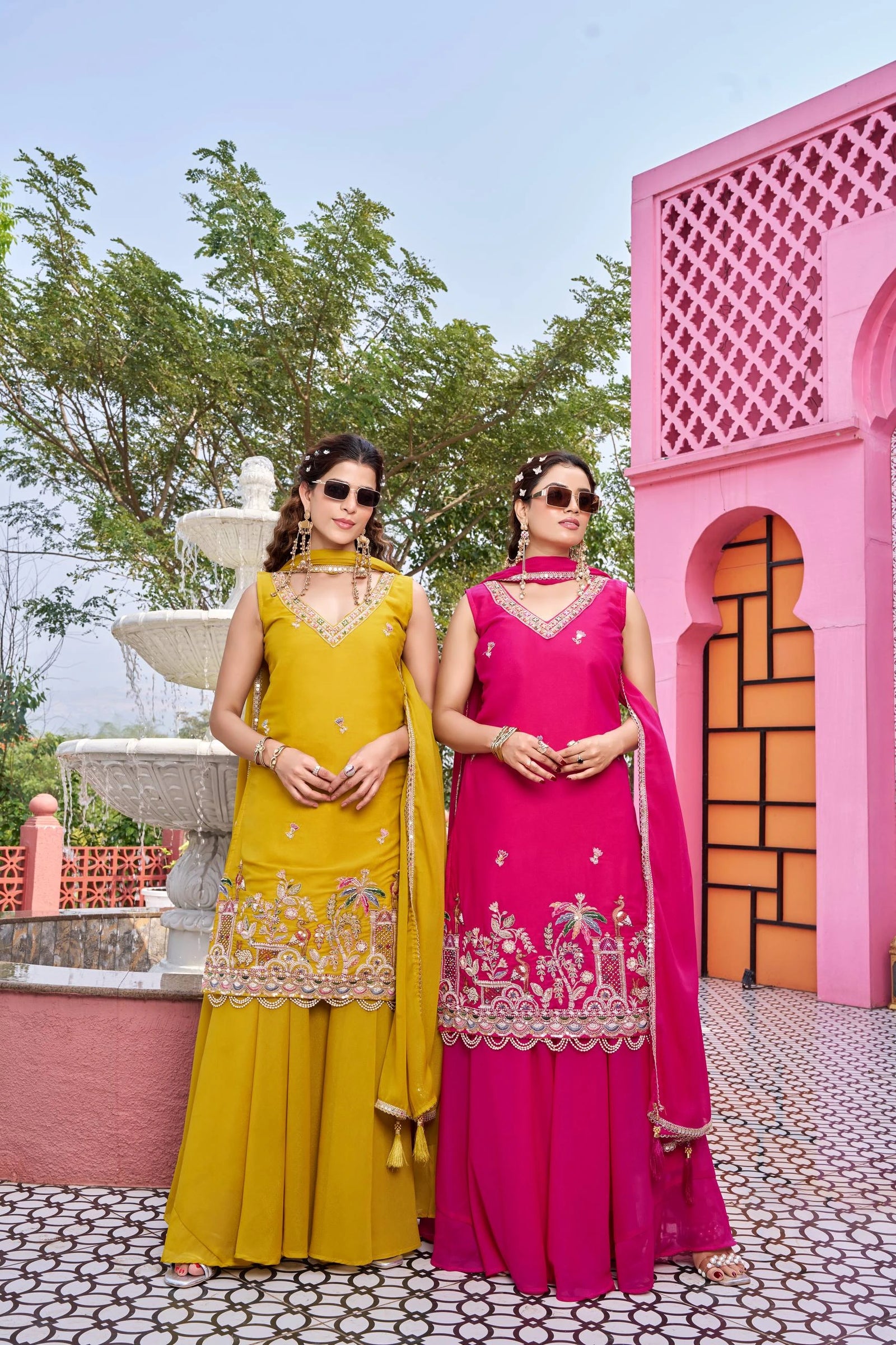 Two women in colorful traditional outfits standing in front of a pink architectural structure.