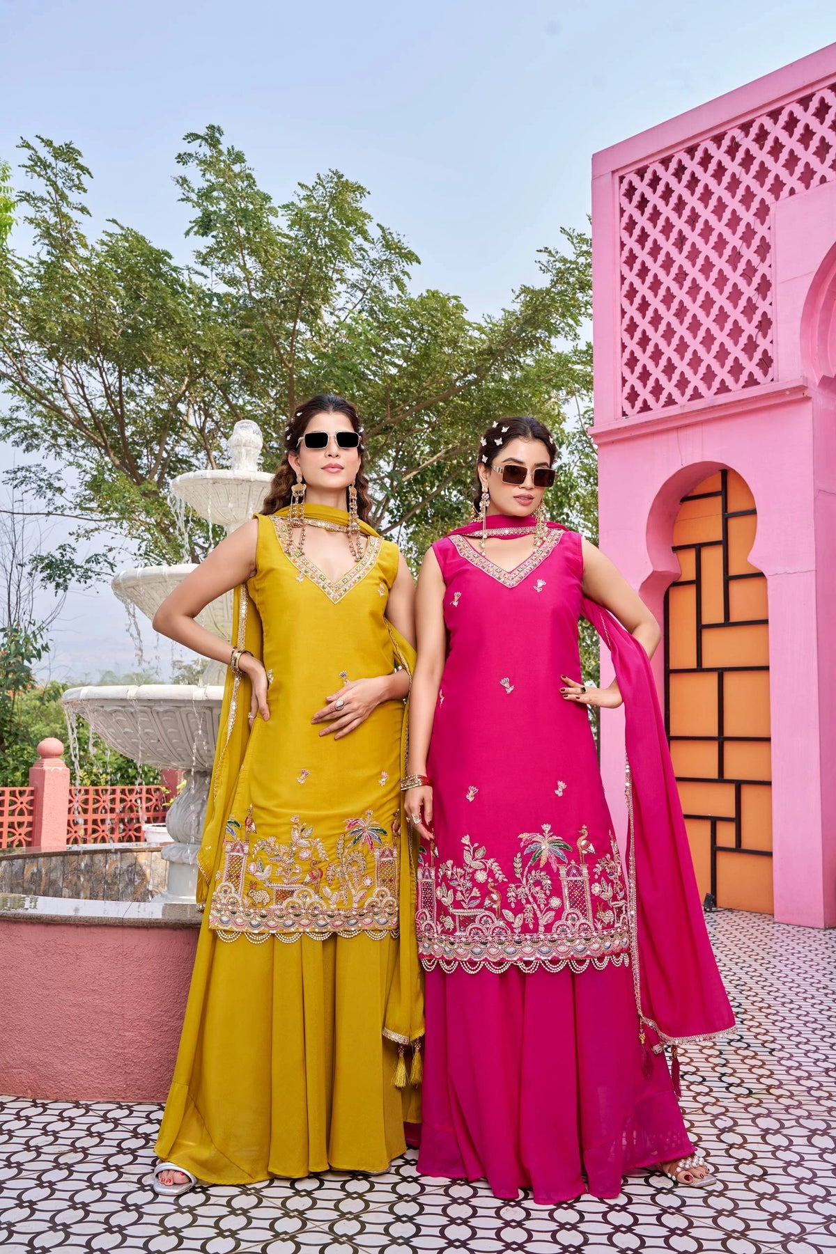 Two women in colorful traditional outfits standing in front of a pink architectural structure.