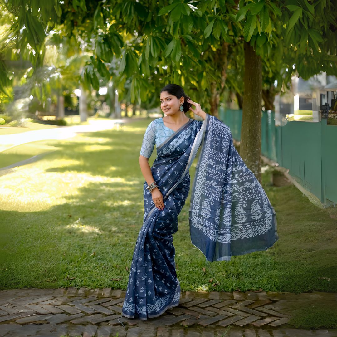 A woman models a striking Navy Blue Soft Cotton Saree featuring an all-over digital block print of traditional white motifs. She wears a light blue patterned blouse and silver jhumka earrings, posing on a path in a green outdoor setting.
