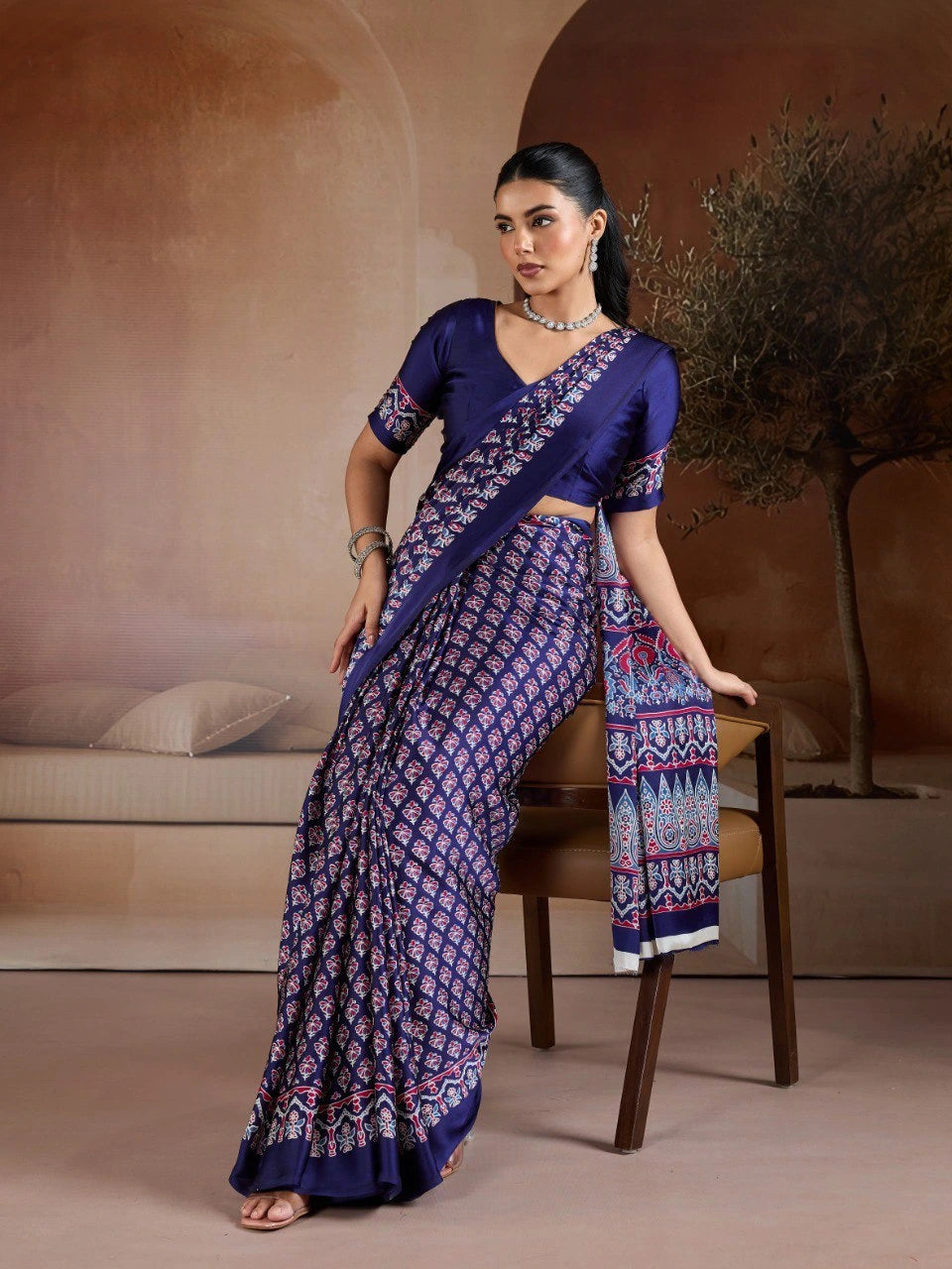 Woman wearing a blue saree with a pattern, sitting on a chair against a neutral background.