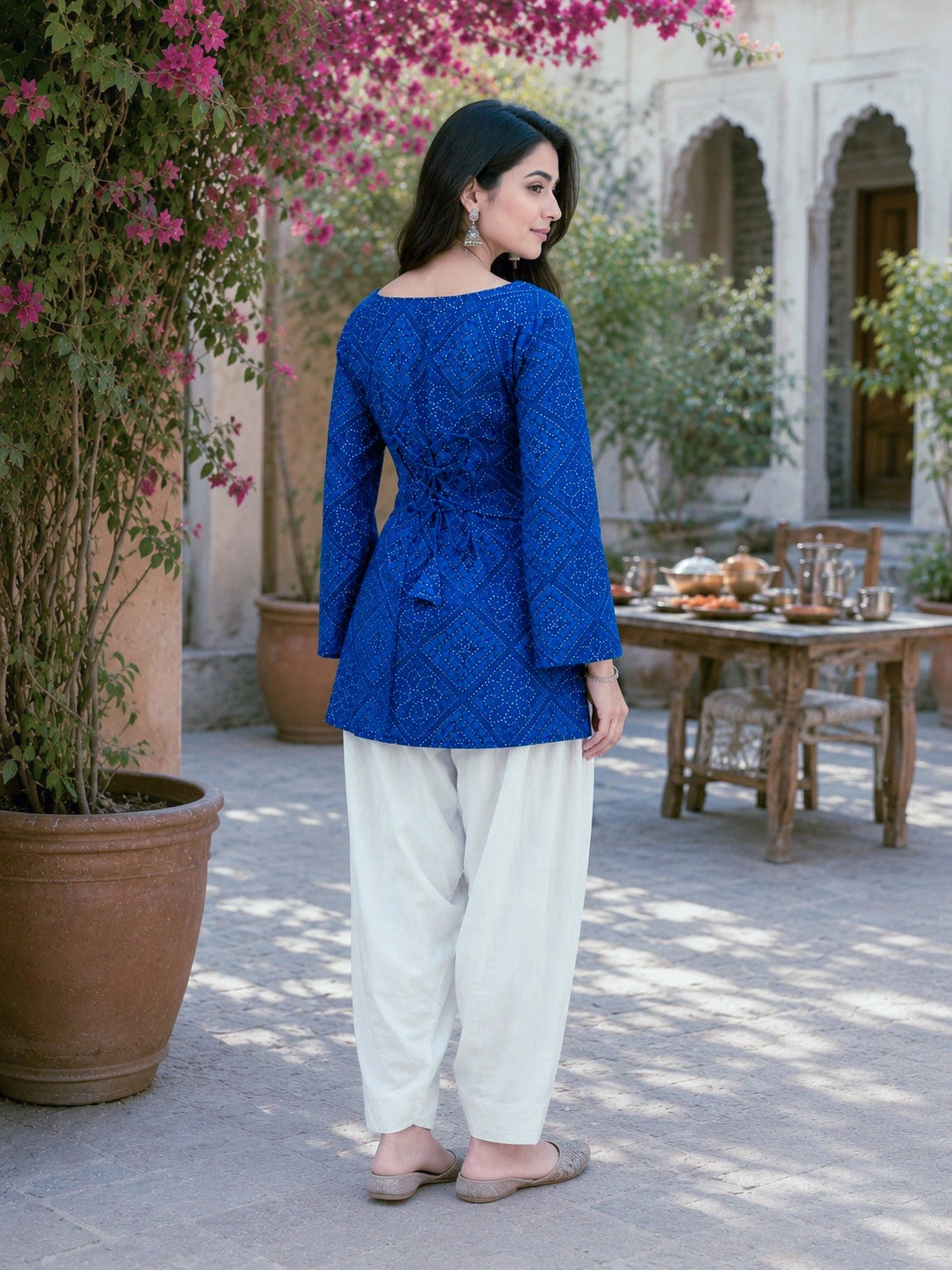 Woman in a blue top and white pants standing in an outdoor setting with plants and a table.
