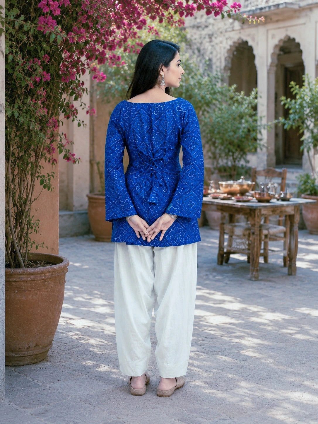 Woman in a blue top and white pants standing in a courtyard with plants and a table in the background.