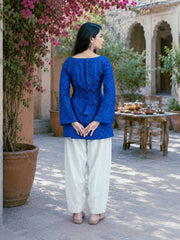 Woman in a blue top and white pants standing in a courtyard with plants and a table in the background.
