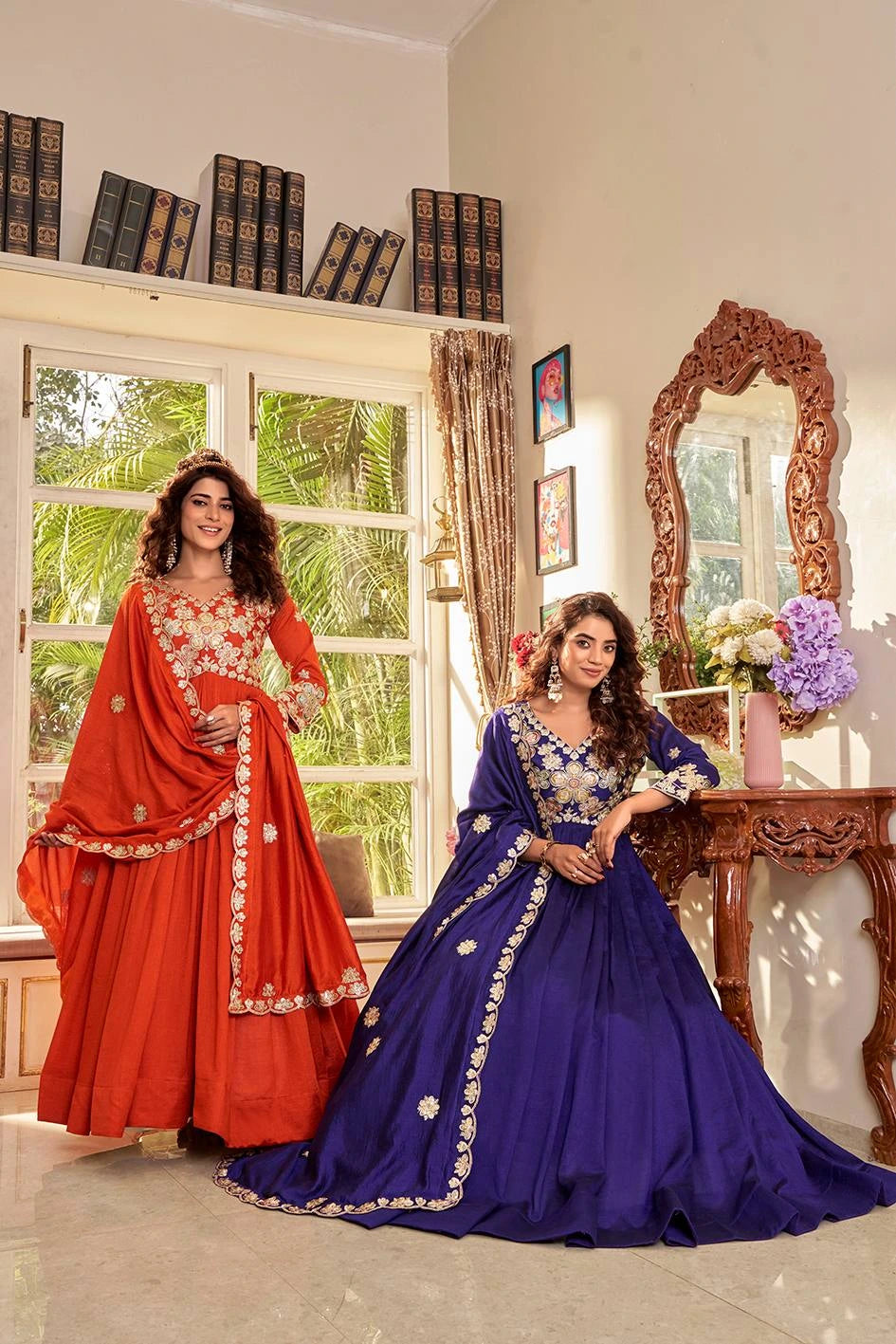Two women in traditional red and blue dresses standing in a room with a decorative mirror and books.