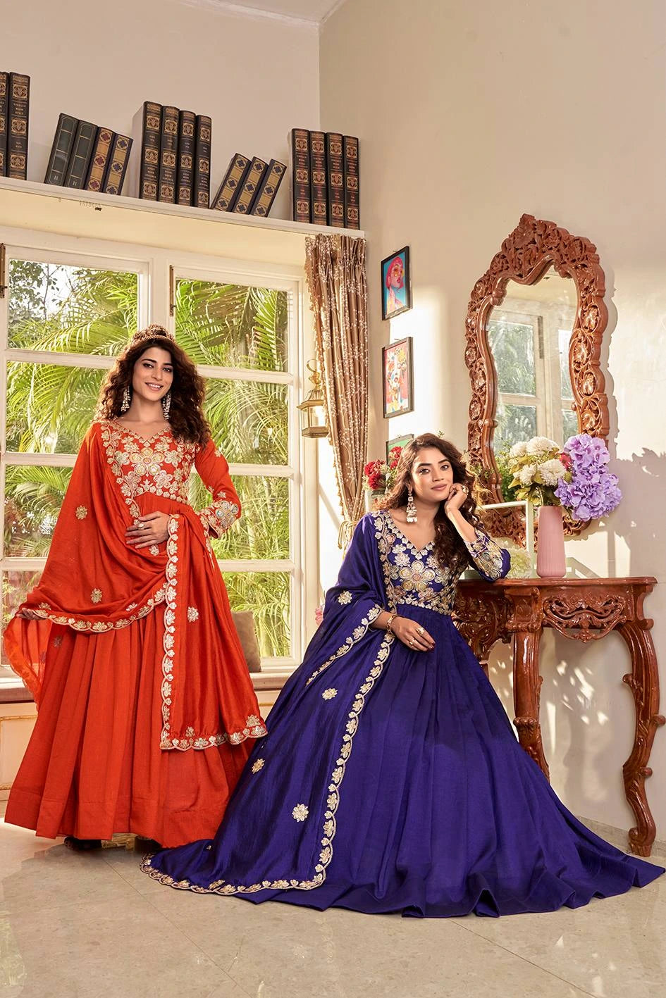 Two women in traditional red and blue dresses standing in a room with a window and decorative mirror.