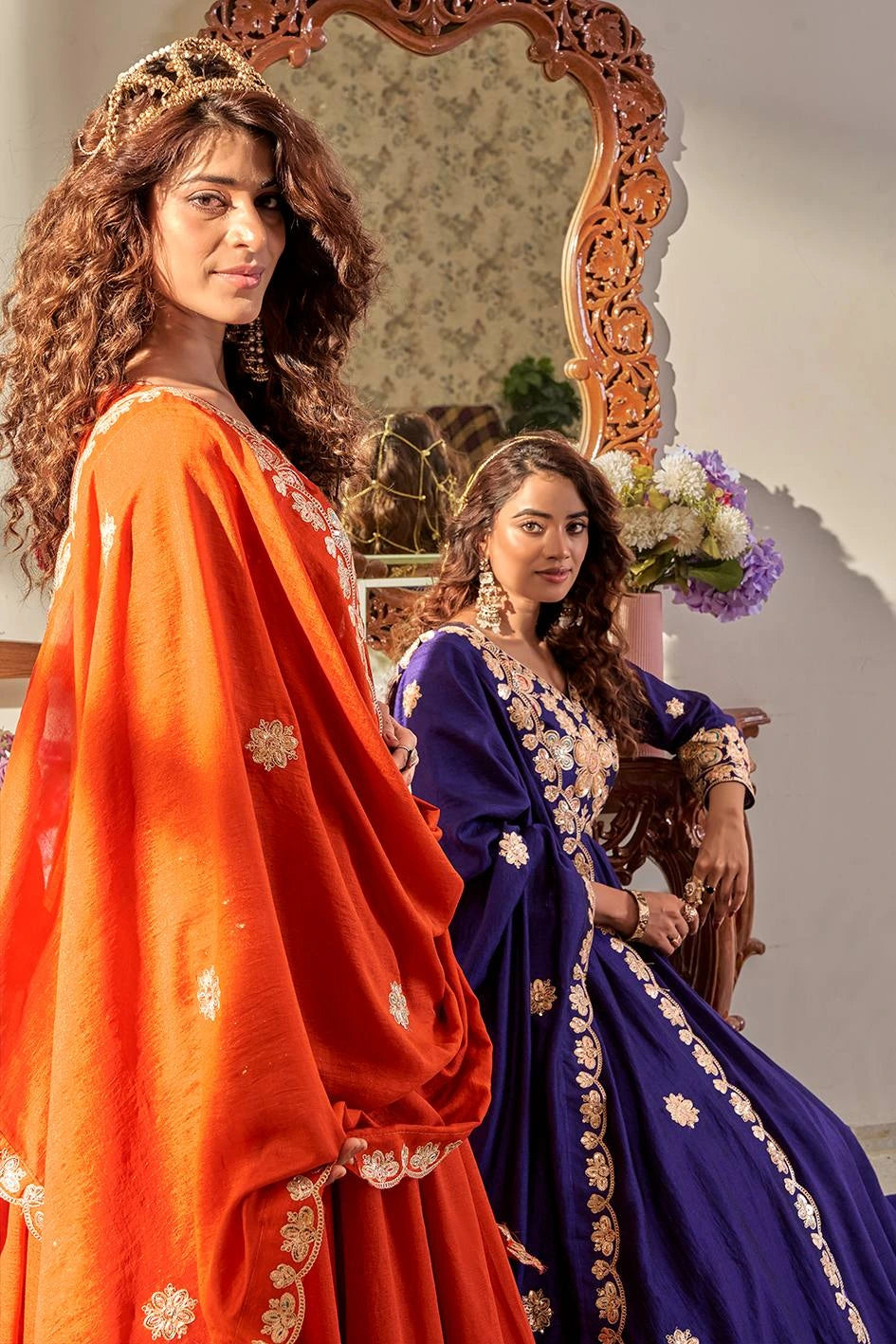 Two women in traditional orange and blue sarees standing in front of a decorative mirror.
