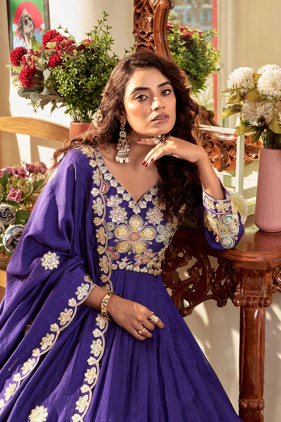 Woman in a blue embroidered traditional outfit sitting in a decorated room with flowers and a mirror.