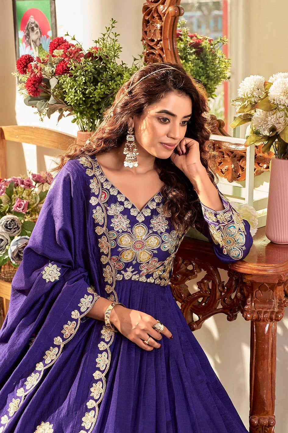 Woman in a blue embroidered traditional outfit sitting in a decorated room with flowers and furniture.