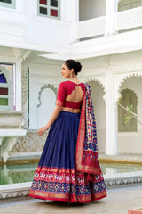 A woman wearing a traditional Indian lehenga choli outfit stands in a courtyard. The outfit features a navy blue pleated skirt with a red and gold patterned border, a red short-sleeve blouse with an open back, and a matching navy blue dupatta with red and gold patterns.