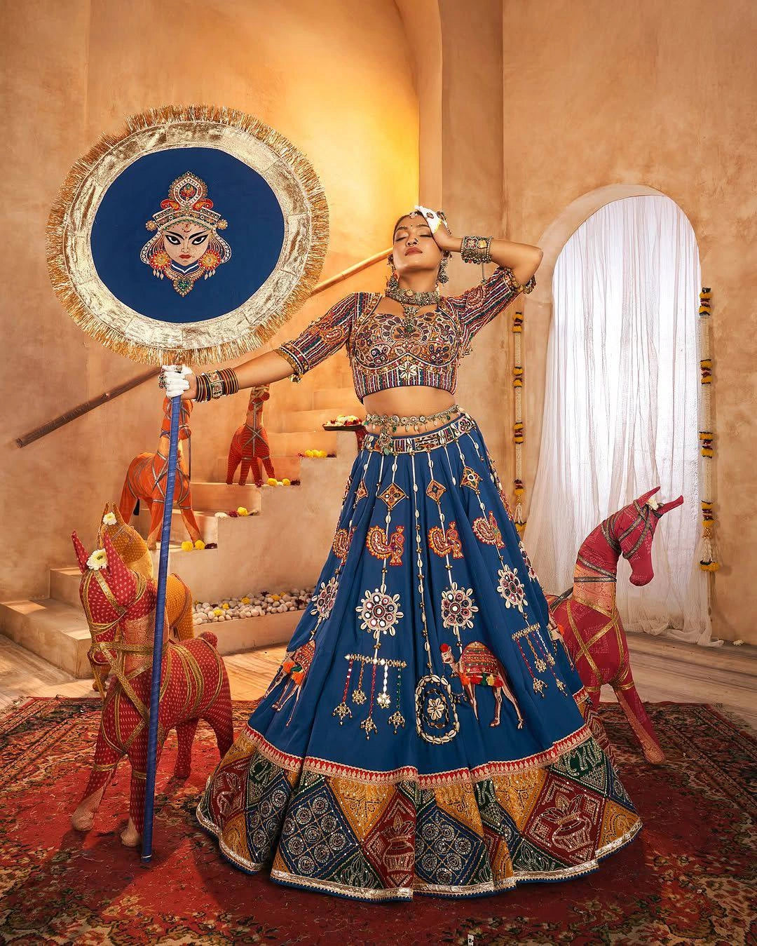 Woman in traditional blue embroidered outfit with decorative elements in a decorated room.