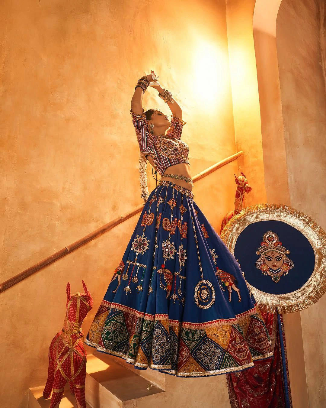 Woman in traditional blue and multicolor lehenga with intricate patterns, standing against a warm-toned wall.