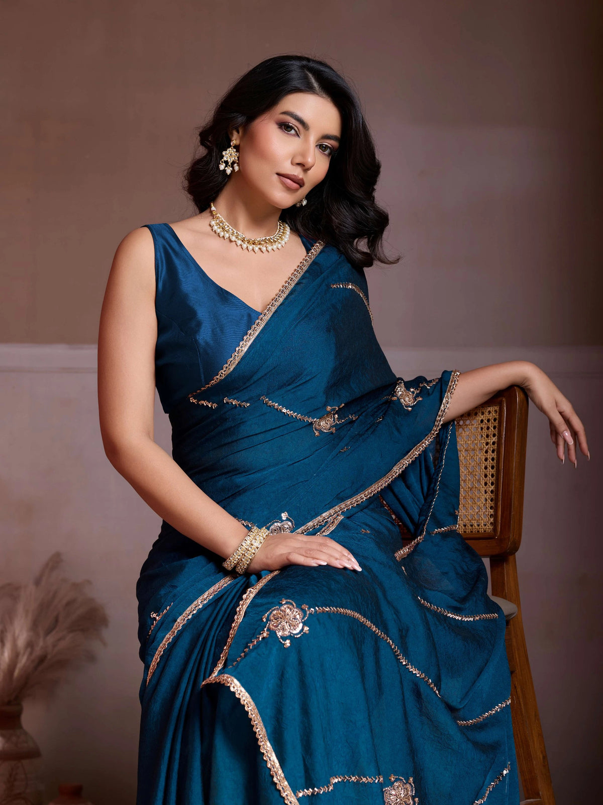 Woman in a blue saree with gold embroidery sitting on a chair against a plain background