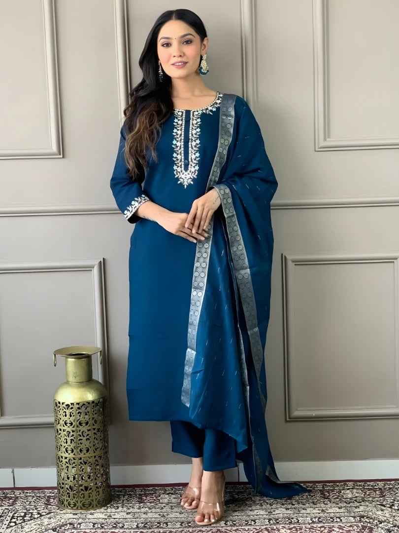 Woman in a blue embroidered outfit standing against a neutral wall.