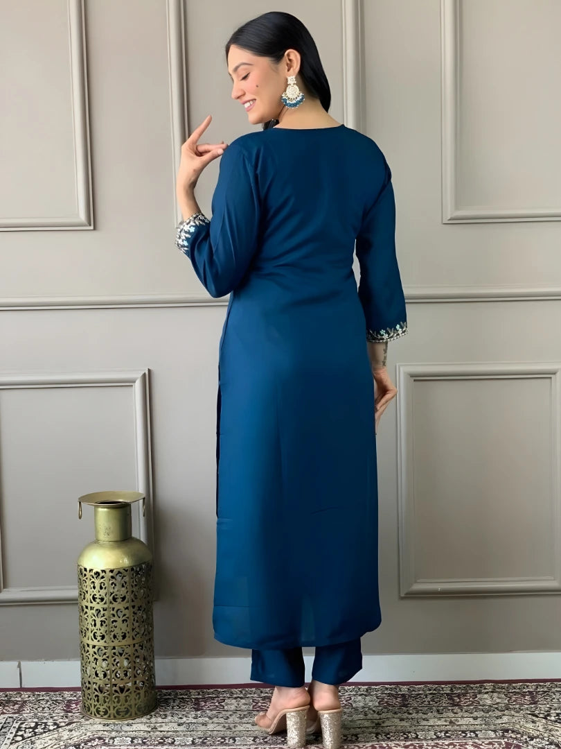 Woman in a blue outfit standing against a gray paneled wall with a decorative vase.