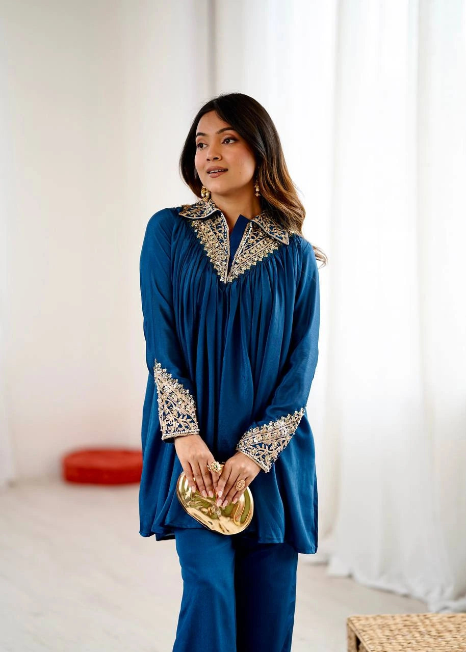 Woman wearing a blue outfit with intricate patterns, holding a gold clutch, in a bright room.