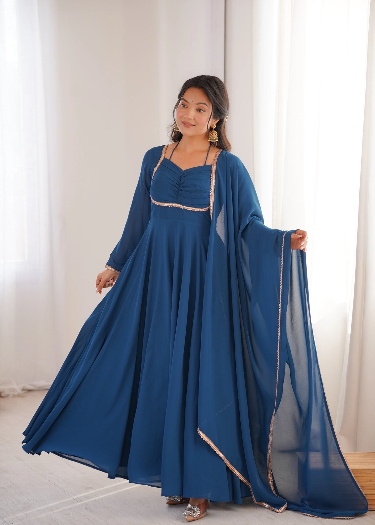 Woman wearing a long blue dress with a sheer overlay in a bright room.