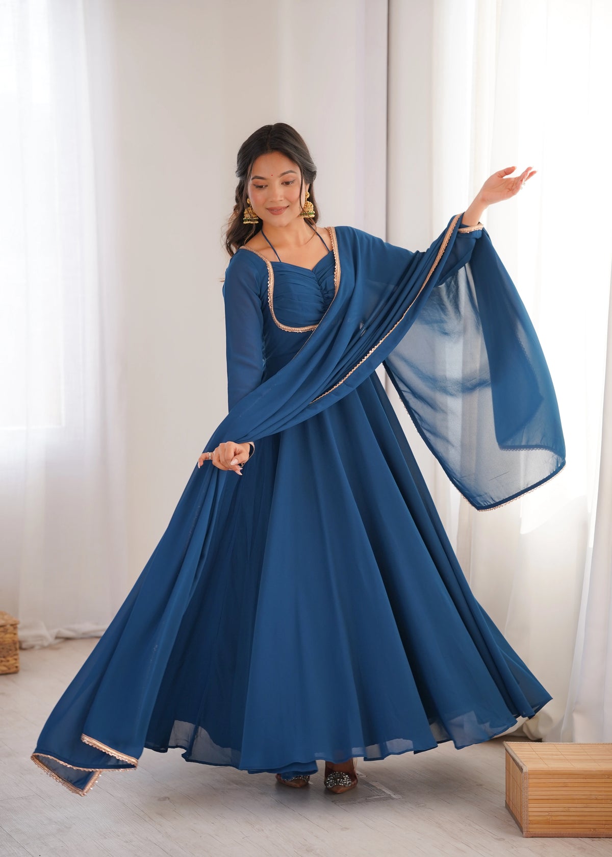 Woman wearing a blue traditional outfit with a sheer dupatta against a white curtain background