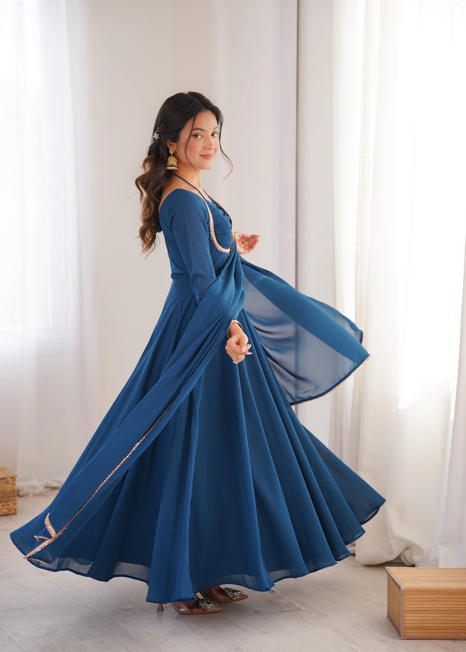 Woman in a blue dress with long sleeves and a flowing skirt standing in a room with white curtains.