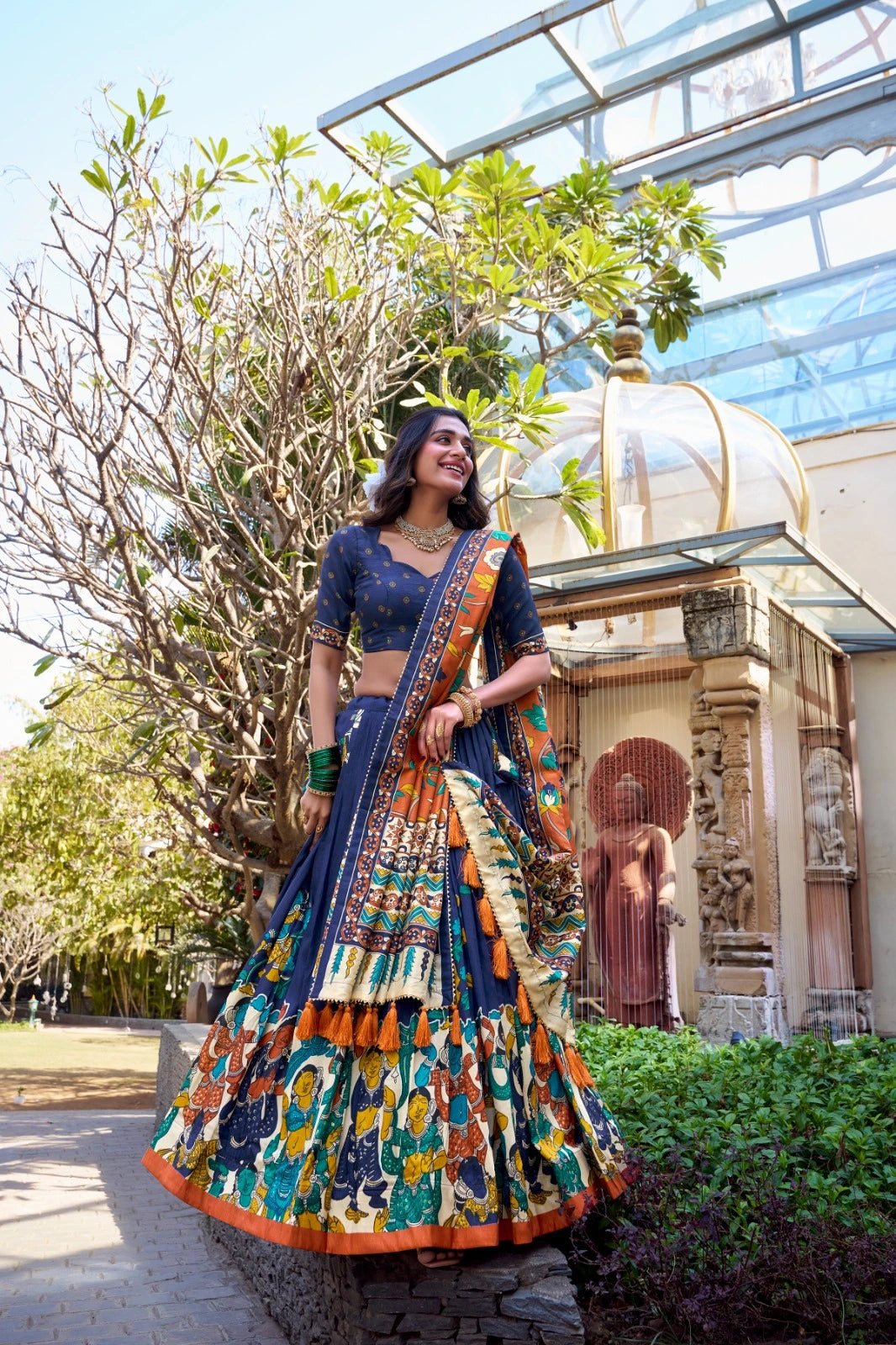 Woman in a traditional outfit standing outdoors with greenery and architectural elements in the background
