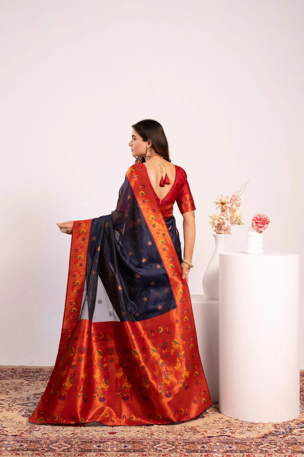 Woman wearing a traditional saree with a red and blue pattern, standing against a plain background.