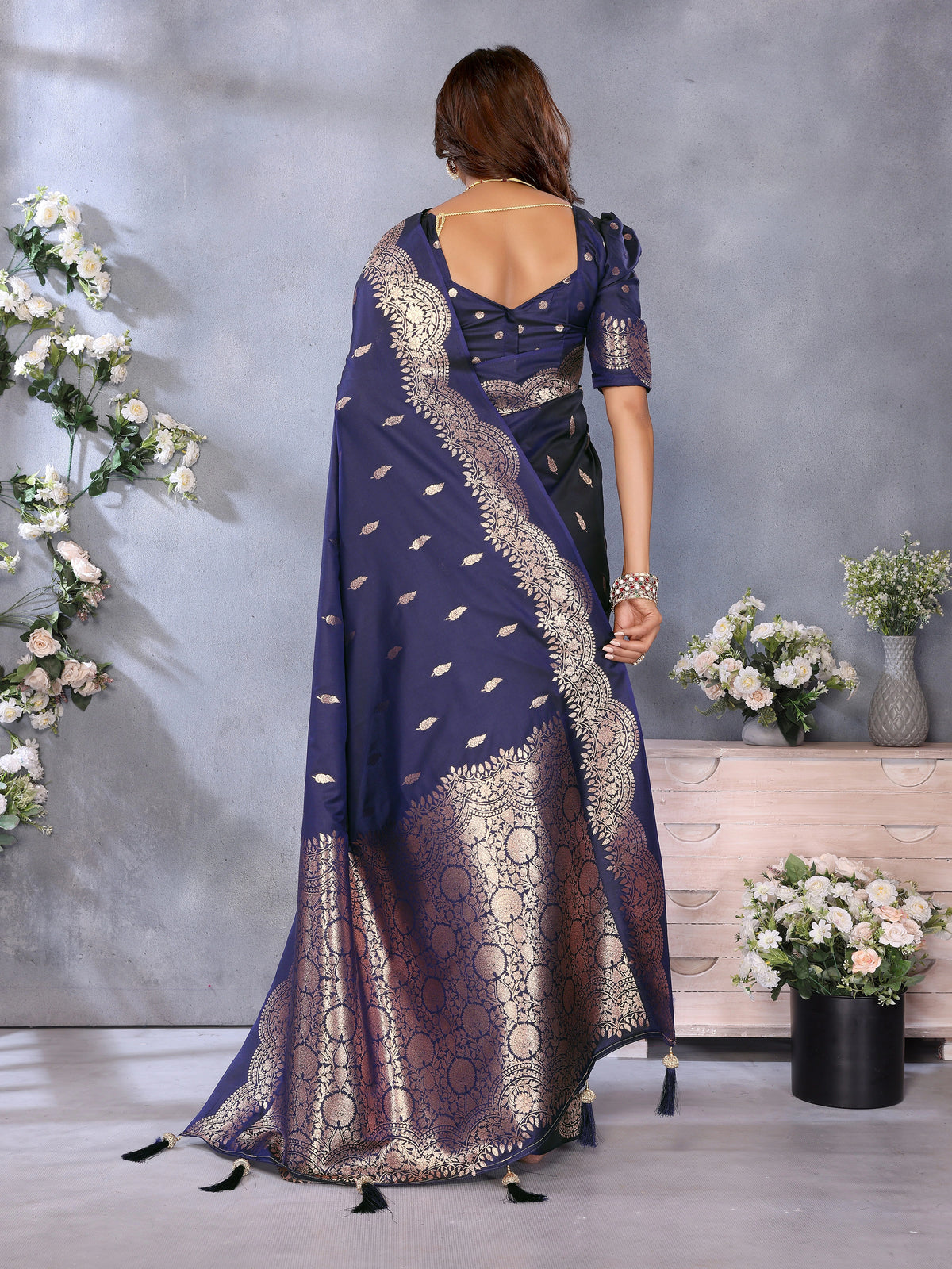 Woman wearing a blue saree with white patterns, standing against a gray wall with floral decorations.