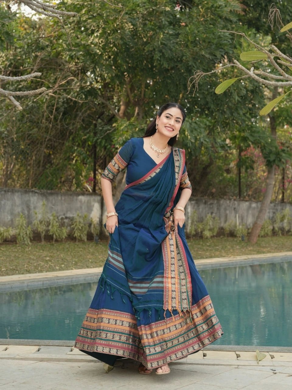 Woman in a blue saree with a colorful border standing by a pool with greenery in the background