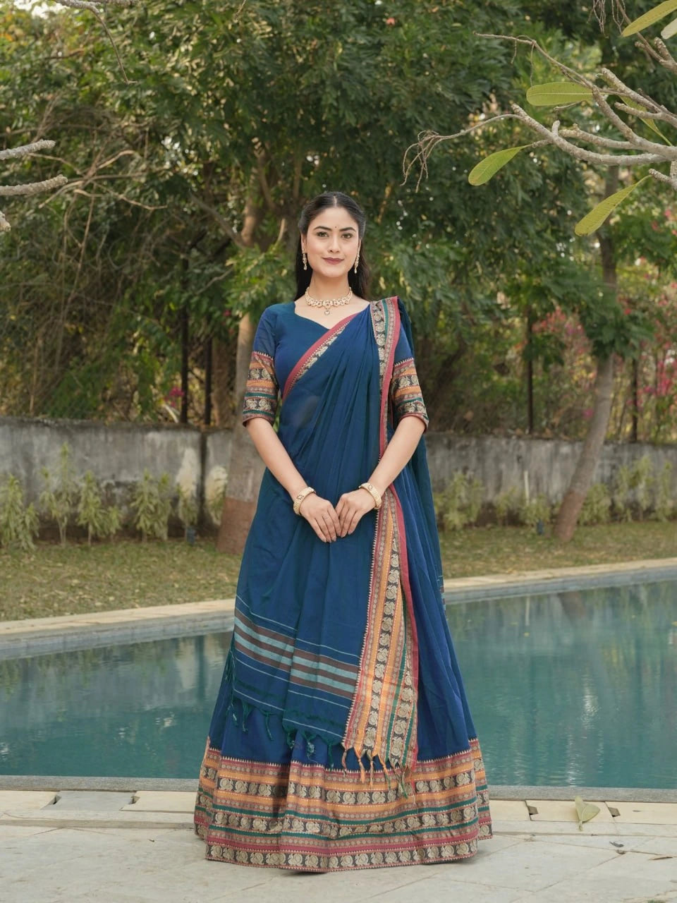 Woman in a blue saree standing by a pool with greenery in the background