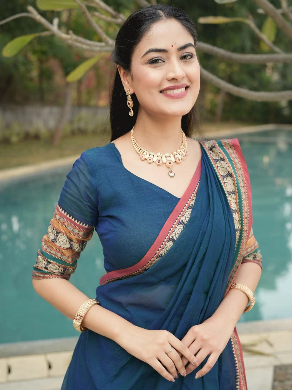Woman in a blue saree with gold jewelry standing by a pool