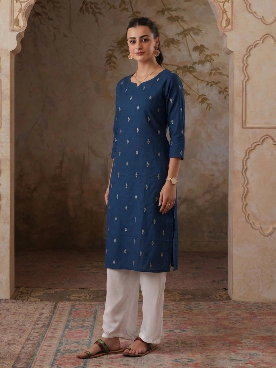 Woman wearing a blue kurta with white patterns and white pants in an ornate room.