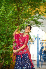 Woman in a pink and blue saree standing among greenery with a blurred background