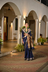 Woman in a traditional blue saree with gold border standing in an archway.