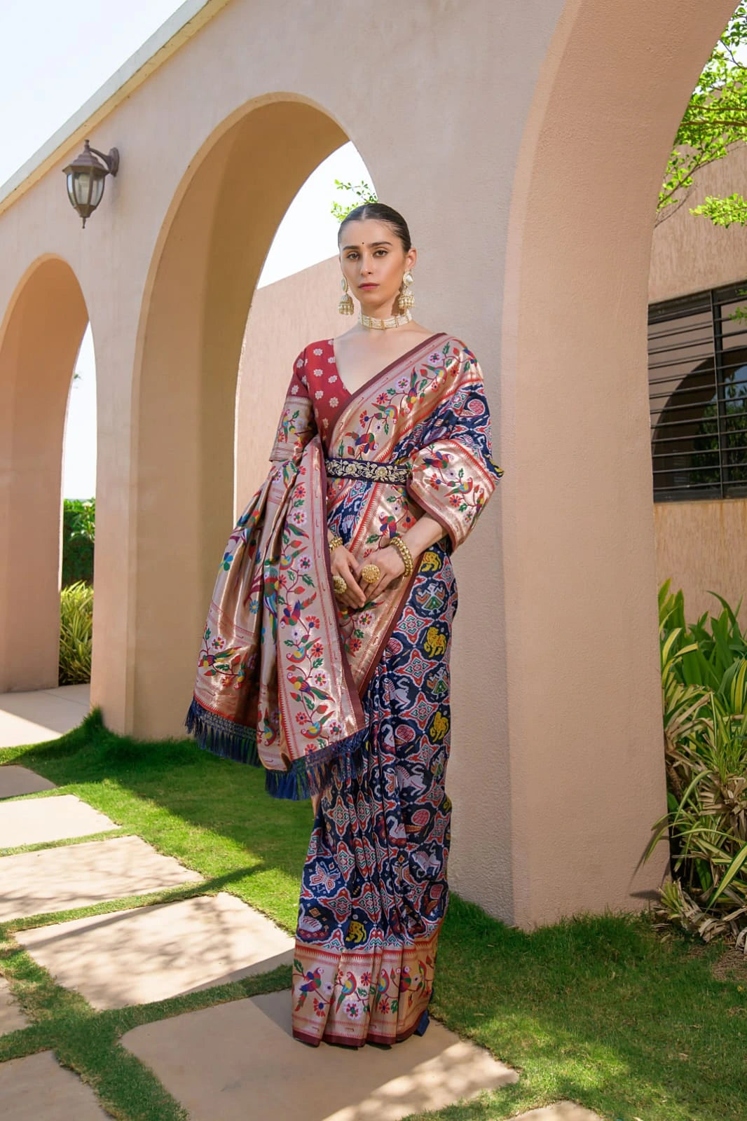 Woman in a colorful saree standing outdoors near a building with arches.