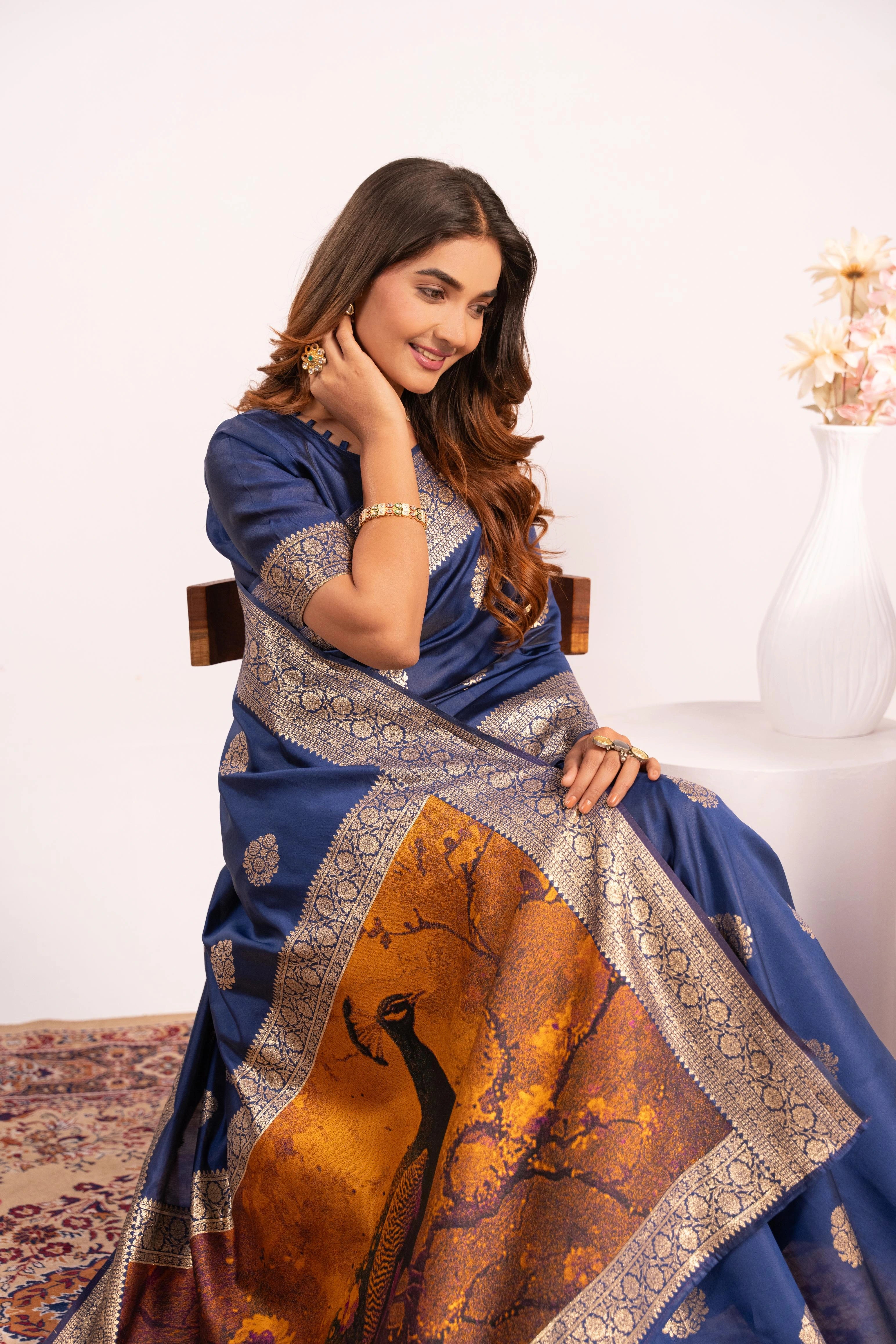 Woman wearing a blue saree with a peacock design, sitting on a white chair.