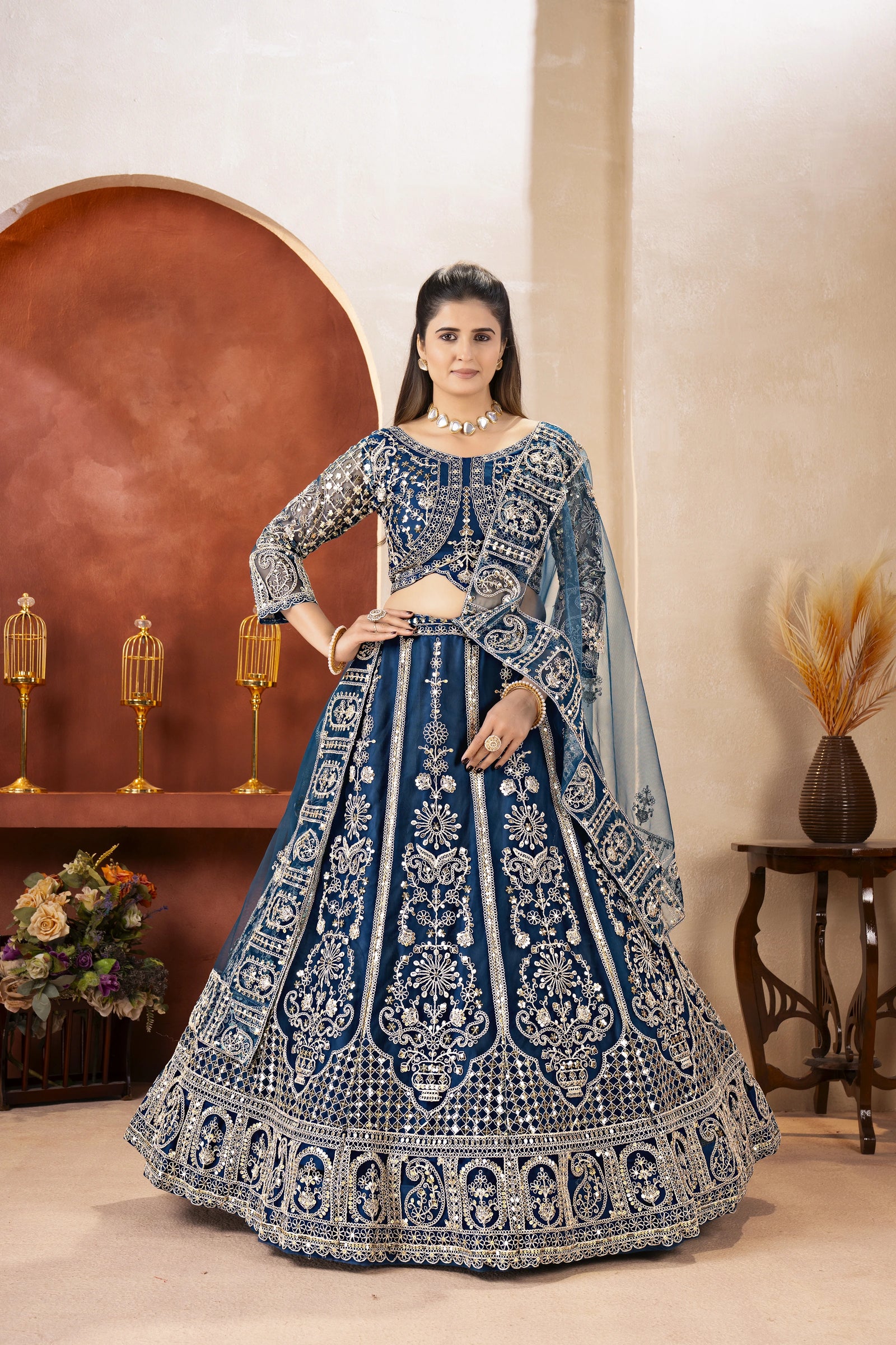 Woman in a blue and silver embroidered traditional outfit standing in a decorated room.
