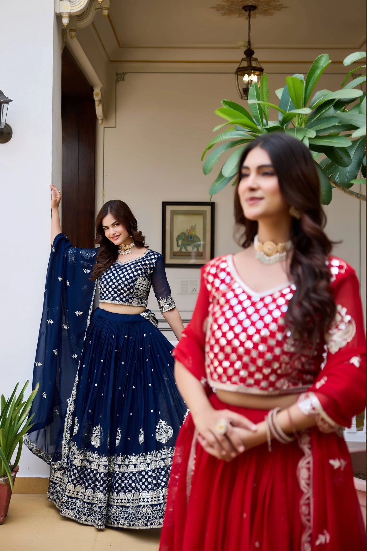 Two women in traditional outfits standing indoors with plants around.