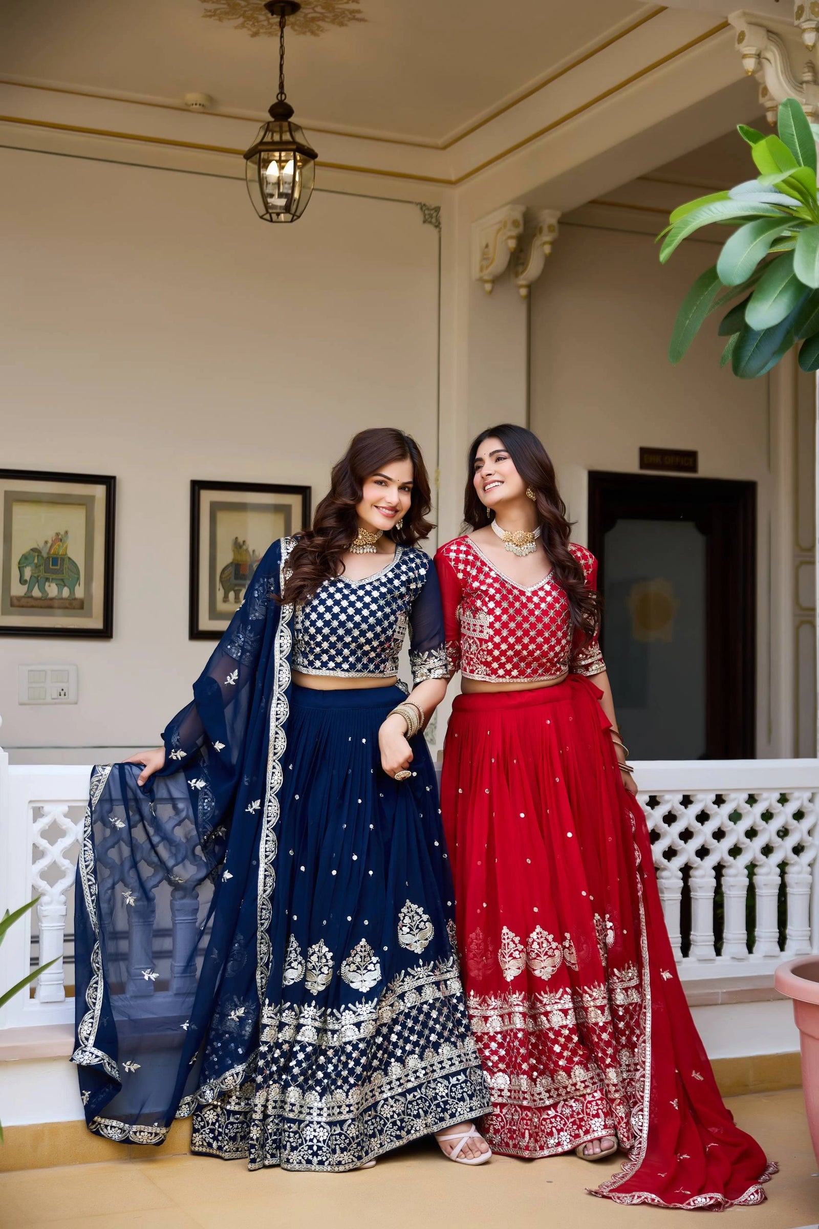Two women in traditional attire standing indoors with decorative elements in the background.