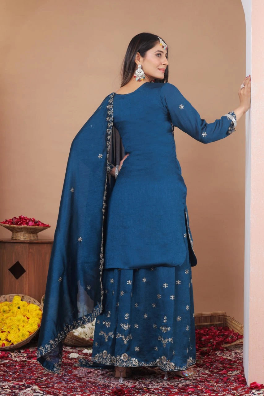 Woman in a blue traditional outfit standing in a decorated room with flowers.